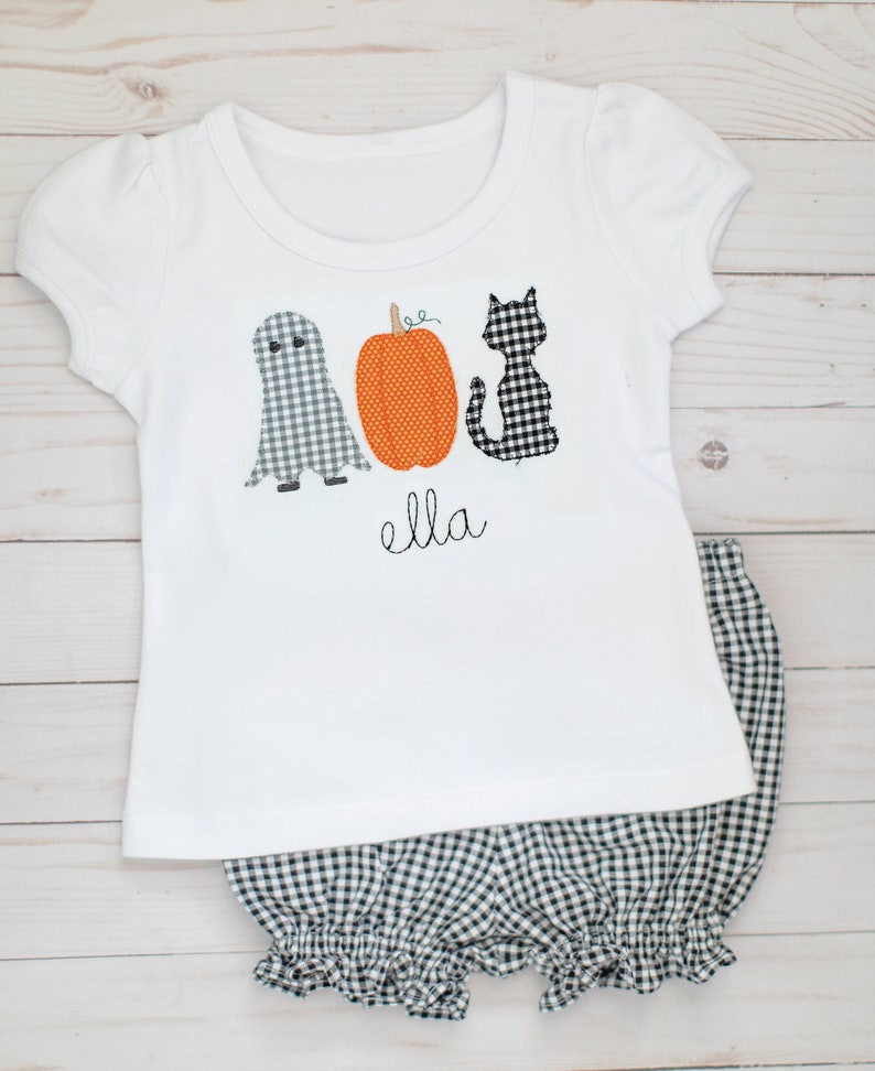 May include: A white short-sleeved t-shirt featuring a ghost, an orange pumpkin, and a black cat in gingham patterns, with the name "ella" embroidered in black script below the designs, paired with black and white gingham ruffled bloomers.