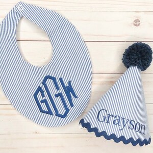 Pode incluir: Um babador listrado azul e branco com um monograma e um chap&eacute;u de festa listrado azul e branco combinando com um pompom azul e o nome Grayson bordado.