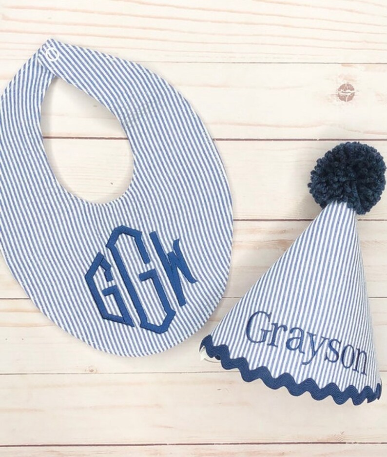 Pode incluir: Um babador listrado azul e branco com um monograma e um chap&eacute;u de festa listrado azul e branco combinando com um pompom azul e o nome Grayson bordado.