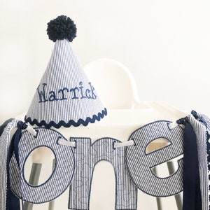 Pode incluir: Uma faixa de tecido listrada azul e branca com a palavra "one" em letras grandes. A faixa &eacute; decorada com uma fita azul e um chap&eacute;u de pompom azul com o nome "Harrick" bordado.