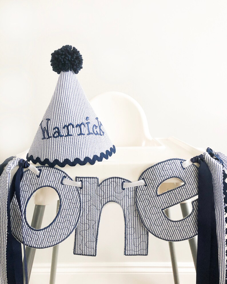 Pode incluir: Uma faixa de tecido listrada azul e branca com a palavra "one" em letras grandes. A faixa &eacute; decorada com uma fita azul e um chap&eacute;u de pompom azul com o nome "Harrick" bordado.