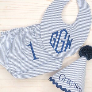 Pode incluir: Um conjunto de roupa de beb&eacute; &agrave;s riscas azuis e brancas com um babador, uma cobertura de fralda e um chap&eacute;u de festa. O babador tem um monograma "GGW" em azul marinho. A cobertura da fralda tem um "1" bordado em azul marinho. O chap&eacute;u de festa tem um pompom azul marinho no topo e o nome "Grayson" bordado no lado.