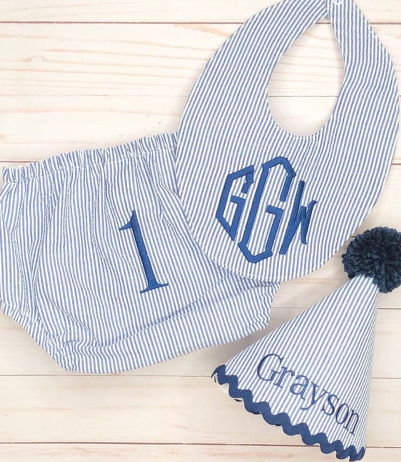 Pode incluir: Um conjunto de roupa de beb&eacute; &agrave;s riscas azuis e brancas com um babador, uma cobertura de fralda e um chap&eacute;u de festa. O babador tem um monograma "GGW" em azul marinho. A cobertura da fralda tem um "1" bordado em azul marinho. O chap&eacute;u de festa tem um pompom azul marinho no topo e o nome "Grayson" bordado no lado.