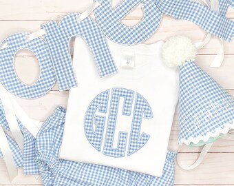 May include: A baby outfit set with a white t-shirt featuring a blue monogram, blue gingham bloomers, and a matching party hat. A banner spelling "ONE" in blue gingham letters is also included. The set is designed for a birthday celebration.