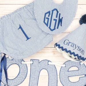 Pode incluir: Uma faixa de tecido listrada azul e branca com a palavra "one" em letras azuis grandes. A faixa &eacute; decorada com costuras brancas. Tamb&eacute;m est&aacute; inclu&iacute;do um babador listrado azul e branco com o monograma "GGW". Tamb&eacute;m est&aacute; inclu&iacute;do um chap&eacute;u listrado azul e branco com o nome "Grayson" bordado.