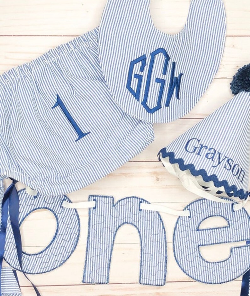 Pode incluir: Uma faixa de tecido listrada azul e branca com a palavra "one" em letras azuis grandes. A faixa &eacute; decorada com costuras brancas. Tamb&eacute;m est&aacute; inclu&iacute;do um babador listrado azul e branco com o monograma "GGW". Tamb&eacute;m est&aacute; inclu&iacute;do um chap&eacute;u listrado azul e branco com o nome "Grayson" bordado.