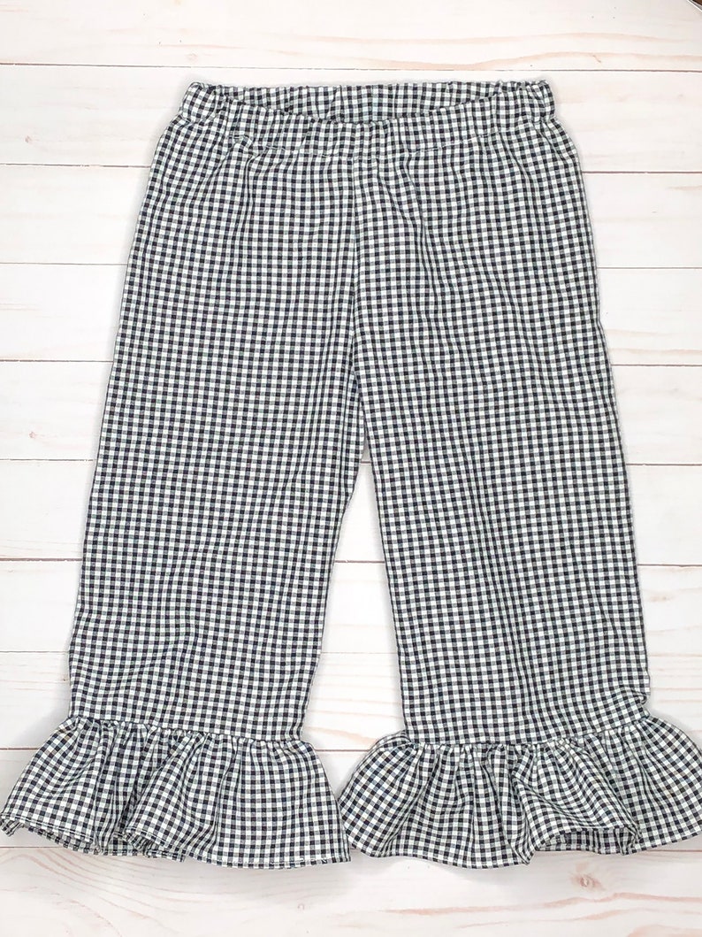 May include: A pair of pants featuring a black and white gingham check pattern. The pants have an elasticized waistband and wide legs that finish with a gathered ruffle detail at the bottom hem, creating a distinct flared silhouette throughout.