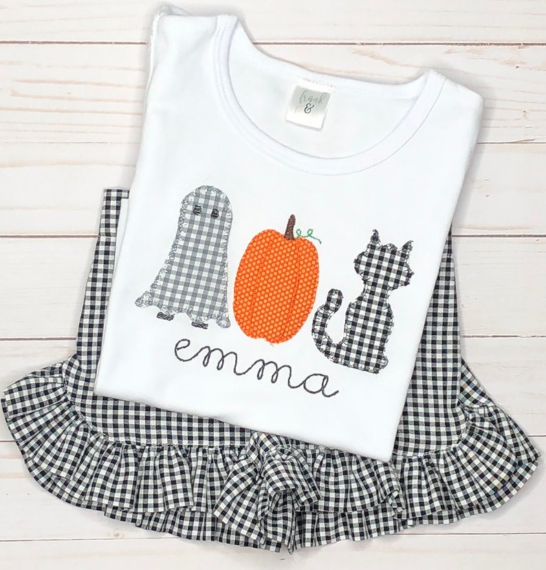 May include: A white t-shirt featuring an applique of a grey gingham ghost, an orange polka-dot pumpkin, and a black gingham cat, with the name emma embroidered below. The shirt is layered over a black and white gingham skirt with a ruffled hem.