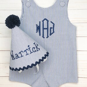 Pode incluir: Um macac&atilde;o listrado azul e branco com um monograma azul marinho "WAG" bordado na frente. Um chap&eacute;u de festa listrado azul e branco combinando com um pompom azul marinho e o nome "Harrick" bordado na lateral.