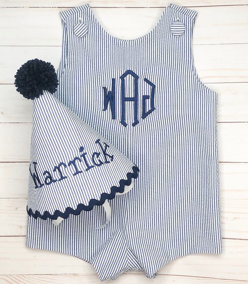 Pode incluir: Um macac&atilde;o listrado azul e branco com um monograma azul marinho "WAG" bordado na frente. Um chap&eacute;u de festa listrado azul e branco combinando com um pompom azul marinho e o nome "Harrick" bordado na lateral.