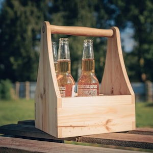 May include: A wooden beer caddy with a handle, holding three bottles of beer. The caddy is made of light-colored wood and sits on a wooden surface. The bottles are filled with a golden liquid, and the background is blurred.