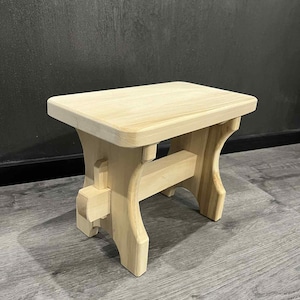May include: A small, unfinished wooden step stool. The stool has a rectangular top and curved legs. The wood is light in color and appears to be new. The stool is designed for use as a step or a small seat.