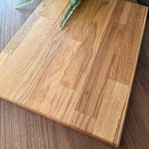 May include: A rectangular, light brown wooden cutting board with a natural wood grain pattern. The board is made of several pieces of wood joined together. A green plant is in the background.