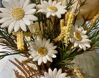 May include: A bouquet of artificial flowers in a white vase. The flowers are white daisies with yellow centers and brown leaves. The vase is decorated with a brown and white pattern.