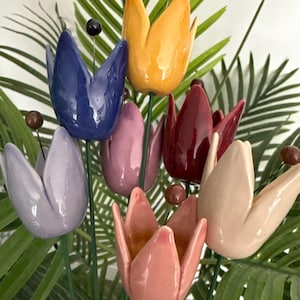 May include: A collection of seven ceramic tulip-shaped garden stakes in various glossy colours including yellow, blue, lavender, pink, red, and cream. Each flower is mounted on a thin green metal stem and features a small brown bead at the centre.