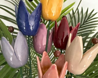 May include: A collection of seven ceramic tulip-shaped garden stakes in various glossy colors including yellow, blue, lavender, pink, red, and cream. Each flower is mounted on a thin green metal stem and features a small brown bead at the center.