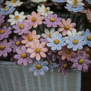 May include: A display of ceramic daisy flowers in a white wicker basket. The flowers are in shades of white, pink, purple, and blue with yellow centres. The text "ceramic flowers" is at the bottom.