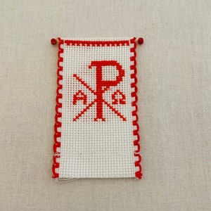 May include: A white cross stitch with a red embroidered Chi Rho symbol, a Christian symbol representing Christ. The cross stitch is framed with a red scalloped border.