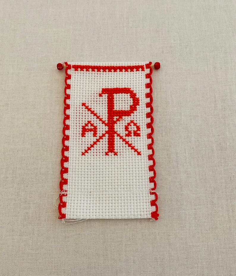 May include: A white cross stitch with a red embroidered Chi Rho symbol, a Christian symbol representing Christ. The cross stitch is framed with a red scalloped border.