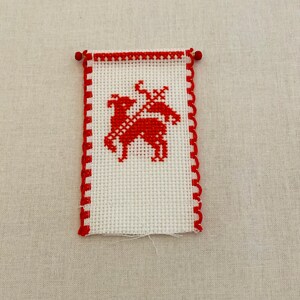 May include: A small white cross-stitch with a red border and a red deer design in the center.