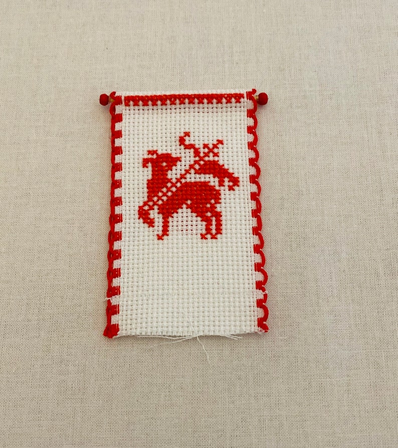 May include: A small white cross-stitch with a red border and a red deer design in the center.