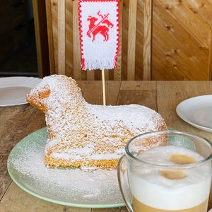 May include: A white cake shaped like a lamb, decorated with powdered sugar, sits on a green plate. A small red and white flag with a lamb design is stuck into the cake. A glass of latte with a thick layer of foam sits on a wooden table next to the cake.