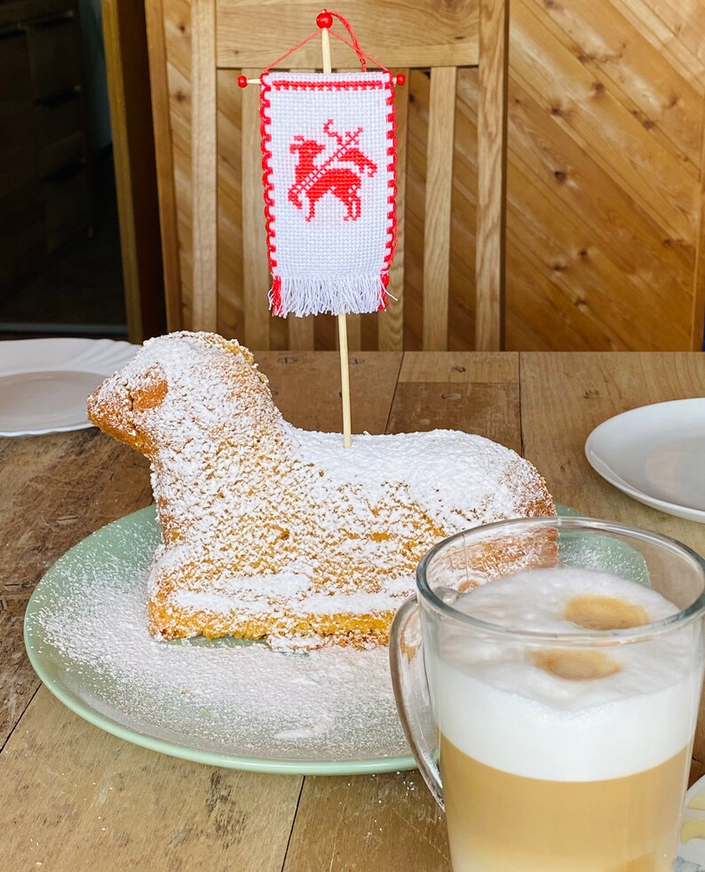 May include: A white cake shaped like a lamb, decorated with powdered sugar, sits on a green plate. A small red and white flag with a lamb design is stuck into the cake. A glass of latte with a thick layer of foam sits on a wooden table next to the cake.