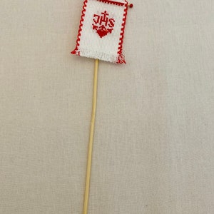 May include: A small white flag with red trim and the letters "IHS" embroidered in red. The flag is attached to a wooden stick.