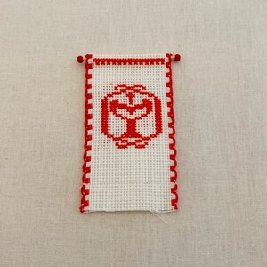 May include: A white cross-stitch banner with a red border and a red cross inside a circle in the center.