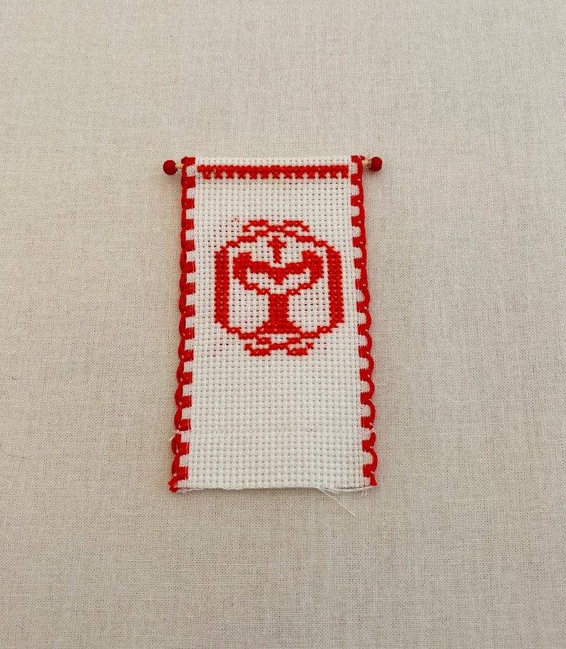 May include: A white cross-stitch banner with a red border and a red cross inside a circle in the center.
