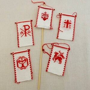 May include: Five small white flags with red trim and red embroidered symbols. The flags are attached to wooden dowels. The symbols include a cross, a heart with the letters "JHS", a chi-rho symbol, a chalice, and a lamb.