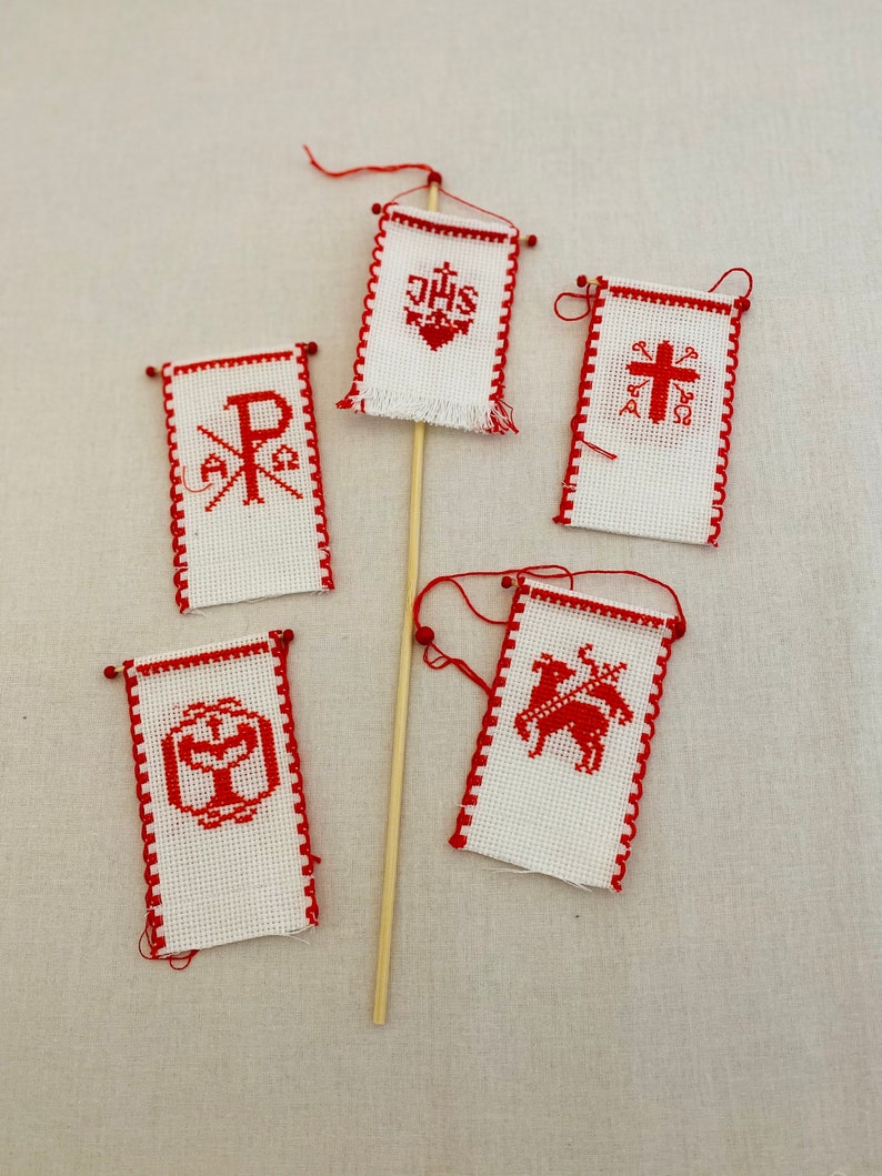 May include: Five small white flags with red trim and red embroidered symbols. The flags are attached to wooden dowels. The symbols include a cross, a heart with the letters "JHS", a chi-rho symbol, a chalice, and a lamb.