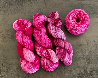 May include: Three skeins of vibrant pink yarn and one ball of matching yarn are displayed. The yarn has a gradient effect, transitioning from deep fuchsia to lighter pink shades. The yarn appears soft and suitable for knitting or crochet projects.