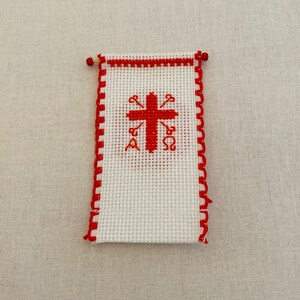 May include: A white cross stitch with a red cross and red Greek letters. The cross stitch is framed with a red border.