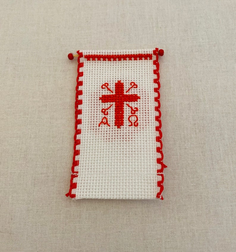 May include: A white cross stitch with a red cross and red Greek letters. The cross stitch is framed with a red border.