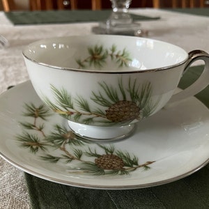 Puede incluir: Una taza de té y un platillo blancos con un diseño de pino y piña. La taza y el platillo tienen un borde dorado.