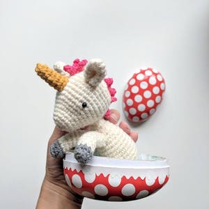 May include: A white crocheted unicorn with a pink mane and a yellow horn sits inside a red and white polka dot Easter egg.