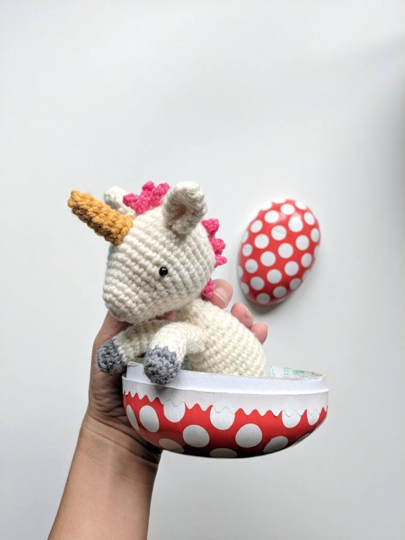 May include: A white crocheted unicorn with a pink mane and a yellow horn sits inside a red and white polka dot Easter egg.