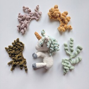 May include: A white crocheted unicorn toy with a light blue mane and horn. The unicorn is surrounded by five crocheted swirls in different colors: pink, orange, green, and brown.