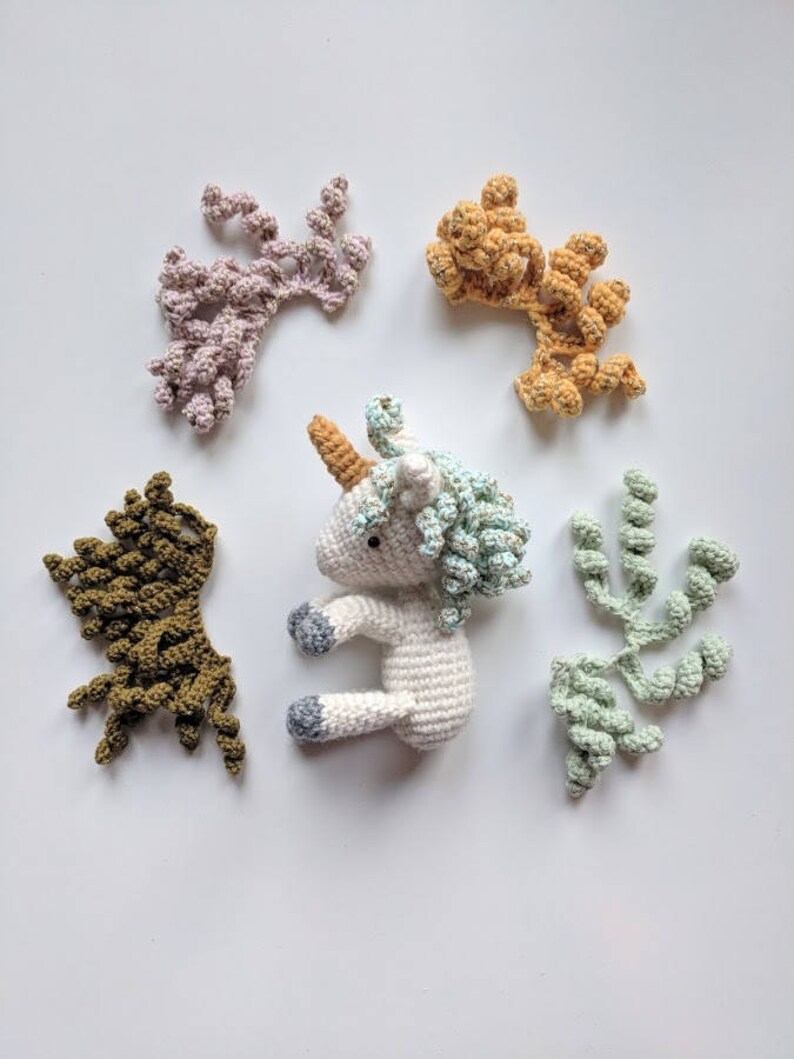 May include: A white crocheted unicorn toy with a light blue mane and horn. The unicorn is surrounded by five crocheted swirls in different colors: pink, orange, green, and brown.