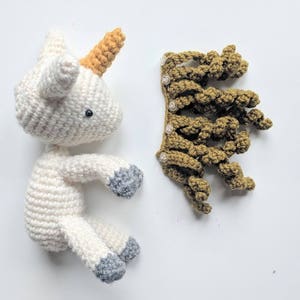 May include: A white crocheted unicorn with a yellow horn and gray hooves. The unicorn is laying on its side with its head turned to the side. A separate crocheted brown mane is also pictured.