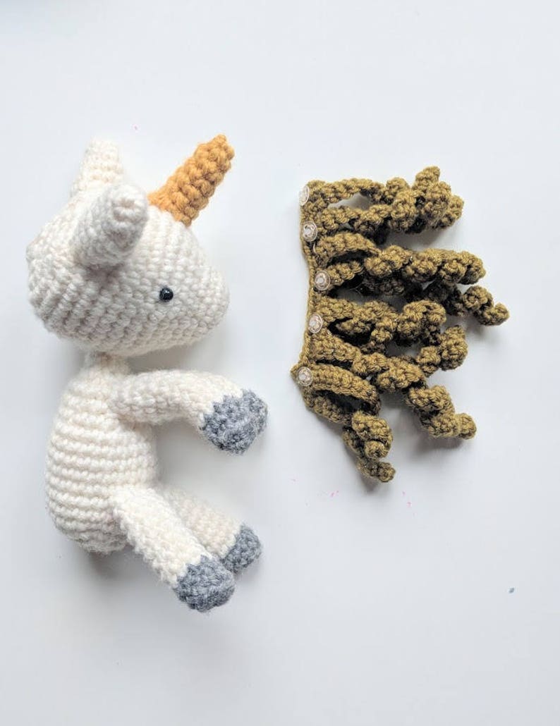 May include: A white crocheted unicorn with a yellow horn and gray hooves. The unicorn is laying on its side with its head turned to the side. A separate crocheted brown mane is also pictured.