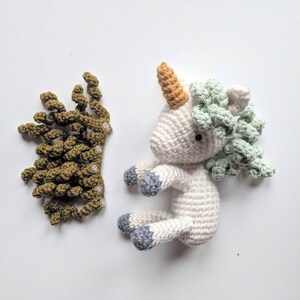 May include: A white crocheted unicorn with a green and light green mane, a light orange horn, and gray hooves. A separate crocheted green crown is also pictured.