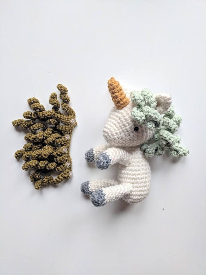 May include: A white crocheted unicorn with a green and light green mane, a light orange horn, and gray hooves. A separate crocheted green crown is also pictured.