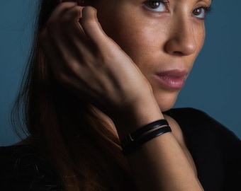 May include: A close-up shot of a person wearing a black leather bracelet with two bands. The bracelet is on the person's wrist. The person is wearing a black top and has long brown hair.