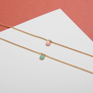May include: Two delicate gold necklaces, each featuring a small, round gemstone pendant. One pendant is a soft pink, the other a light teal. The necklaces rest on a white surface with a coral-colored background.