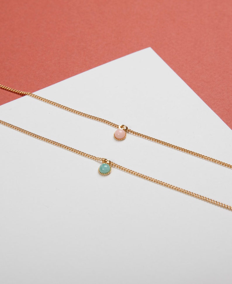 May include: Two delicate gold necklaces, each featuring a small, round gemstone pendant. One pendant is a soft pink, the other a light teal. The necklaces rest on a white surface with a coral-colored background.