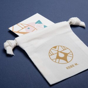 May include: A white canvas drawstring bag with a gold logo and the text "KOBO M." is displayed on a dark blue surface. A white card with a geometric design in pink, teal, and gold is tucked inside the bag.