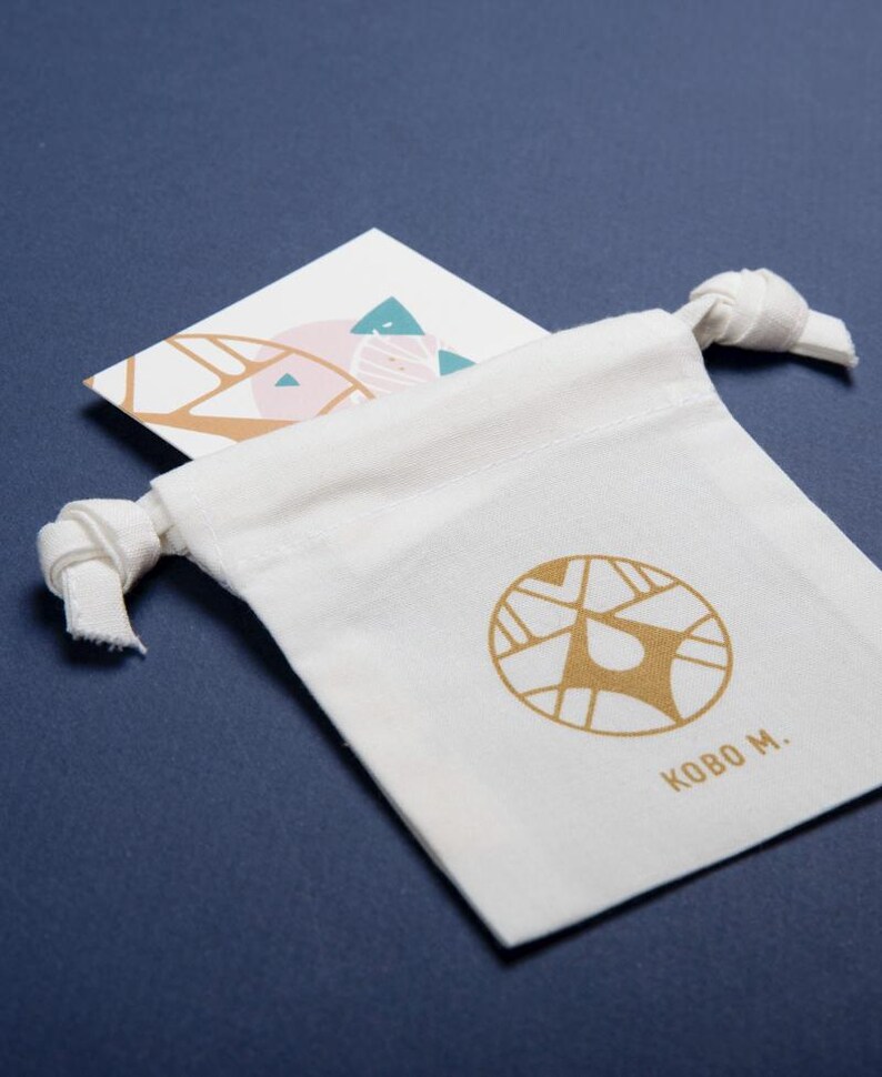 May include: A white canvas drawstring bag with a gold logo and the text "KOBO M." is displayed on a dark blue surface. A white card with a geometric design in pink, teal, and gold is tucked inside the bag.