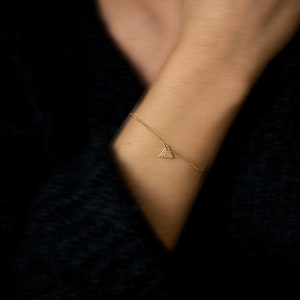 Puede incluir: Una delicada pulsera dorada con un pequeño colgante triangular. El colgante está cubierto de diminutos detalles brillantes. La pulsera se lleva en una muñeca, con un fondo oscuro.
